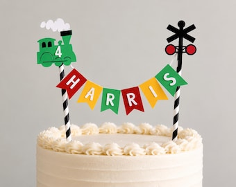 May include: A cake topper featuring a green train engine with the number 4 and a black railroad crossing sign on striped black and white sticks. Between them hangs a bunting banner with red, yellow, and green flags spelling out HARRIS.