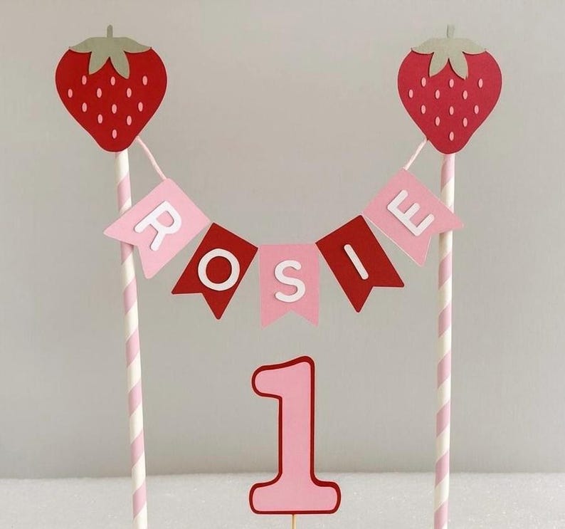 Puede incluir: Adorno para tarta con tem&aacute;tica de fresas en rosa y rojo. Dos fresas rojas grandes con tapas verdes est&aacute;n sobre pajitas a rayas. Una pancarta deletrea "ROSIE" en banderas rosas y rojas. Un n&uacute;mero "1" rosa est&aacute; en la parte inferior.