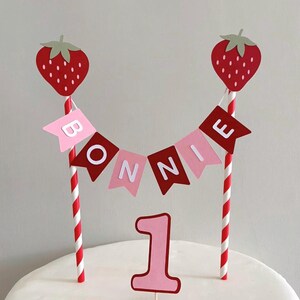 Puede incluir: Adorno para tarta de primer cumplea&ntilde;os. Incluye dos fresas rojas sobre pajitas a rayas rojas y blancas, una guirnalda con el nombre "BONNIE" y el n&uacute;mero "1". La guirnalda y el n&uacute;mero son de color rosa y rojo.