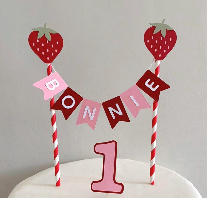 Puede incluir: Adorno para tarta de primer cumplea&ntilde;os. Incluye dos fresas rojas sobre pajitas a rayas rojas y blancas, una guirnalda con el nombre "BONNIE" y el n&uacute;mero "1". La guirnalda y el n&uacute;mero son de color rosa y rojo.