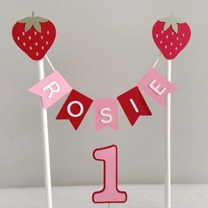 Puede incluir: Adorno para tarta rosa y rojo con el nombre "Rosie" en una pancarta, dos decoraciones de fresas y el n&uacute;mero "1". Las fresas tienen hojas verdes y est&aacute;n unidas a palitos blancos.