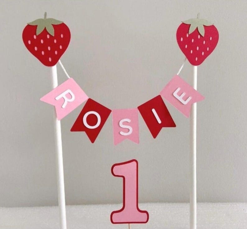 Puede incluir: Adorno para tarta rosa y rojo con el nombre "Rosie" en una pancarta, dos decoraciones de fresas y el n&uacute;mero "1". Las fresas tienen hojas verdes y est&aacute;n unidas a palitos blancos.