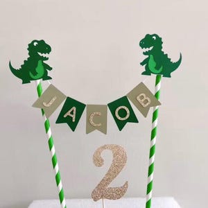 May include: Green dinosaur cake topper with a banner that says "JACOB" and a gold glitter number 2.