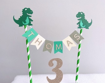 Puede incluir: Topper de pastel de dinosaurio verde con una pancarta que dice "Thomas 3" en letras doradas con purpurina. La pancarta está hecha de papel verde, gris y blanco.