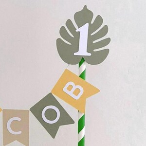 May include: A cake topper featuring a green paper monstera leaf with a white number 1 attached to a green and white striped paper straw. Below the leaf, a small bunting banner displays the letters C, O, and B on individual pennants.