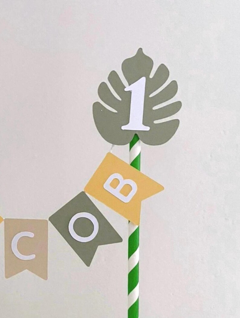 May include: A cake topper featuring a green paper monstera leaf with a white number 1 attached to a green and white striped paper straw. Below the leaf, a small bunting banner displays the letters C, O, and B on individual pennants.
