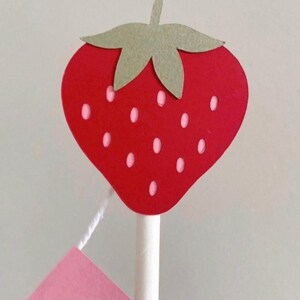 Puede incluir: Decoraci&oacute;n en forma de fresa roja con hojas verdes y peque&ntilde;as semillas blancas. La fresa est&aacute; unida a un palo blanco, con una etiqueta rosa colgando debajo. La etiqueta tiene letras blancas.