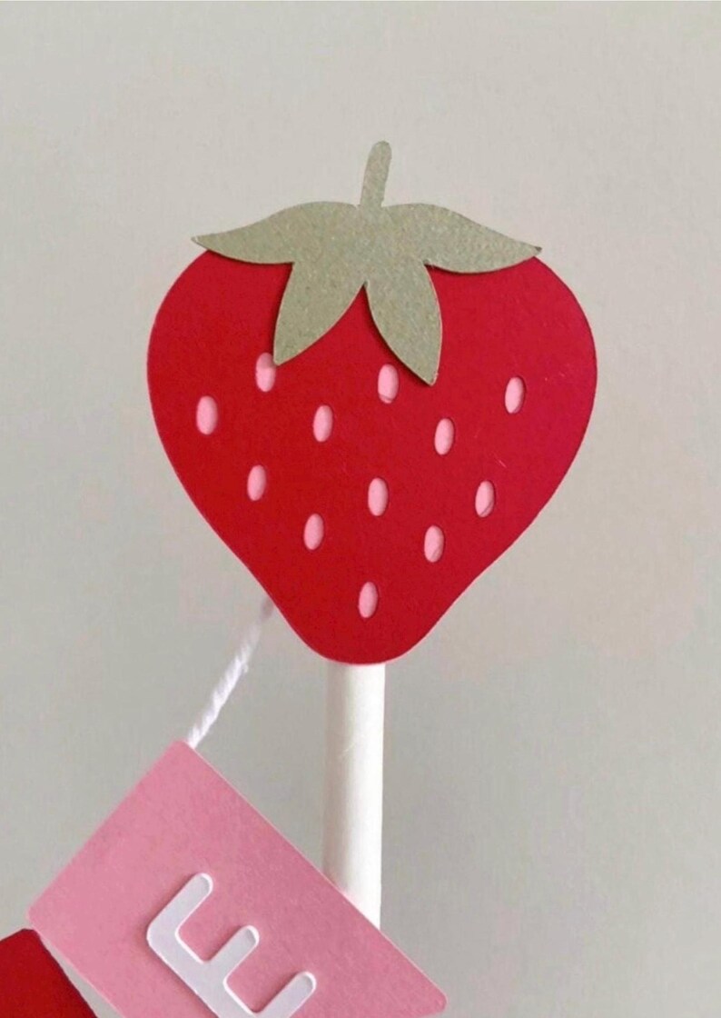 Puede incluir: Decoraci&oacute;n en forma de fresa roja con hojas verdes y peque&ntilde;as semillas blancas. La fresa est&aacute; unida a un palo blanco, con una etiqueta rosa colgando debajo. La etiqueta tiene letras blancas.