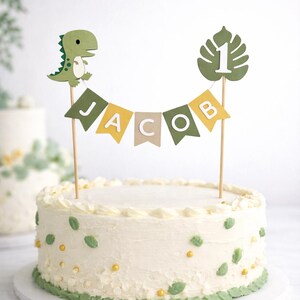 May include: A cake topper set featuring a green dinosaur, a monstera leaf with the number 1, and a bunting banner spelling JACOB in white letters on green, yellow, and beige flags, all mounted on thin wooden sticks atop a white frosted cake.
