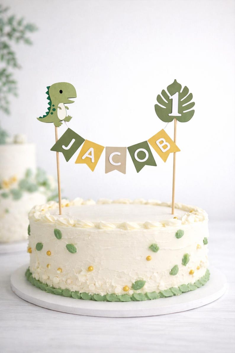 May include: A cake topper set featuring a green dinosaur, a monstera leaf with the number 1, and a bunting banner spelling JACOB in white letters on green, yellow, and beige flags, all mounted on thin wooden sticks atop a white frosted cake.