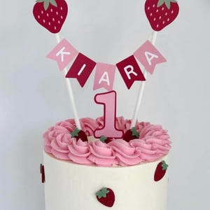 Puede incluir: Un pastel decorado para un primer cumplea&ntilde;os. Presenta glaseado blanco, remolinos rosas, fresas y una pancarta con el nombre "KIARA". El pastel est&aacute; adornado con un n&uacute;mero "1" rosa y adornos de fresa.