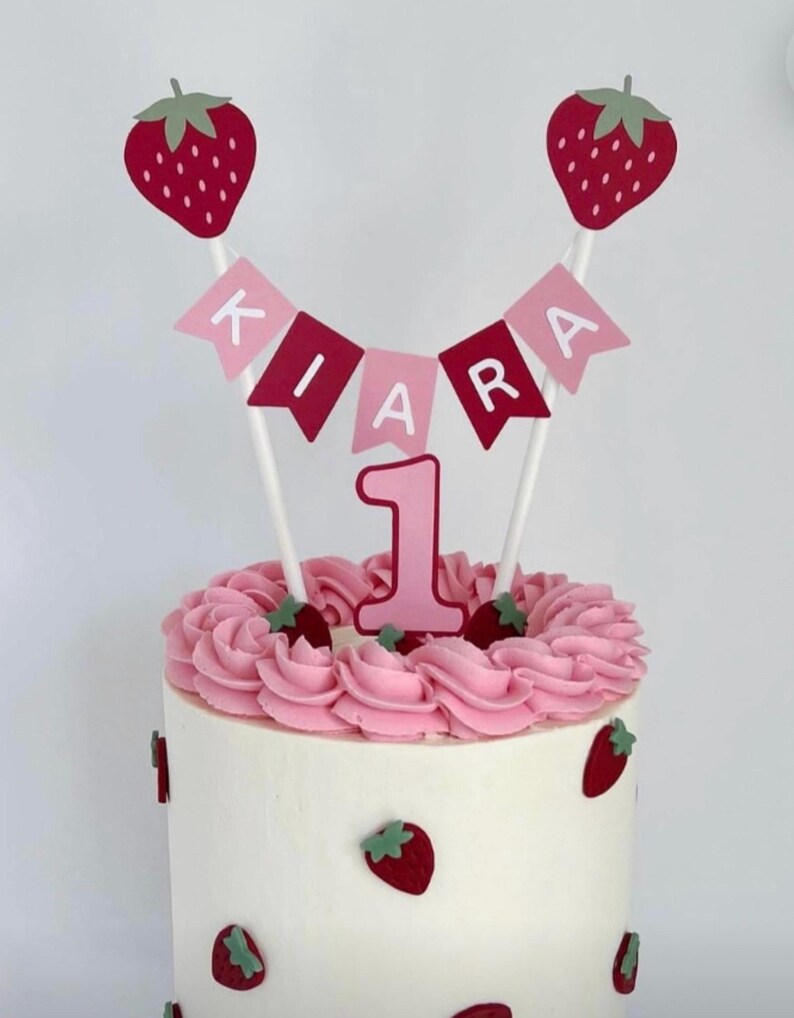 Puede incluir: Un pastel decorado para un primer cumplea&ntilde;os. Presenta glaseado blanco, remolinos rosas, fresas y una pancarta con el nombre "KIARA". El pastel est&aacute; adornado con un n&uacute;mero "1" rosa y adornos de fresa.
