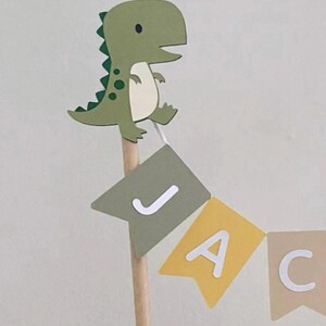 May include: A decorative cake topper featuring a cartoon green dinosaur attached to a wooden stick. Below the dinosaur, a small bunting banner hangs, displaying the letters J, A, and C in white on individual sage green, yellow, and beige pennants.