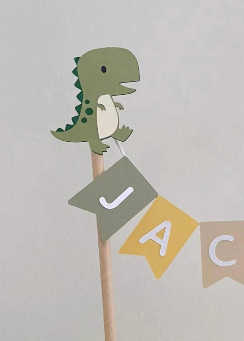 May include: A decorative cake topper featuring a cartoon green dinosaur attached to a wooden stick. Below the dinosaur, a small bunting banner hangs, displaying the letters J, A, and C in white on individual sage green, yellow, and beige pennants.