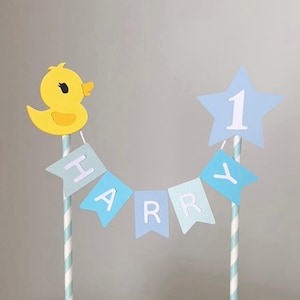 May include: A yellow duck cake topper with a blue and white striped straw. The topper includes a banner that spells out "HARRY" and a blue star with the number "1". The duck has a small orange beak and a black eye.
