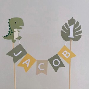 May include: A cake topper featuring a green dinosaur and a monstera leaf with the number 1, connected by a bunting banner with the name JACOB in white letters. The banner consists of alternating olive green, yellow, and beige swallowtail flags.