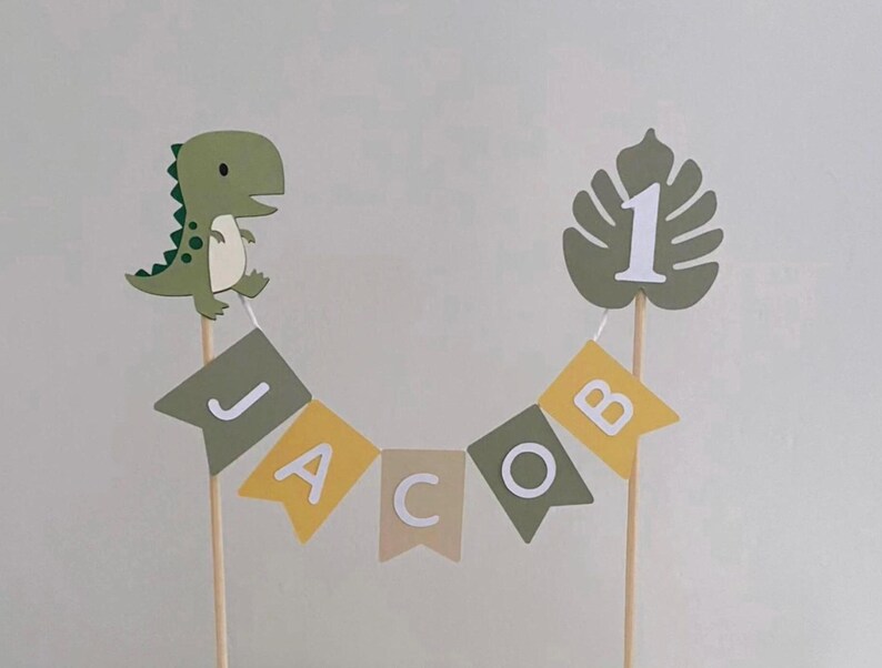 May include: A cake topper featuring a green dinosaur and a monstera leaf with the number 1, connected by a bunting banner with the name JACOB in white letters. The banner consists of alternating olive green, yellow, and beige swallowtail flags.