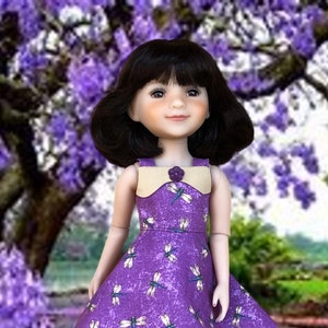 Puede incluir: Un vestido morado con estampado de libélulas y un cuello beige. El vestido tiene un cierre de botón en el escote. La muñeca lleva el vestido y tiene el pelo negro y los ojos marrones.
