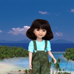 Puede incluir: Una muñeca con una camisa de manga corta azul claro y un mono verde con estampado floral. La muñeca tiene el pelo castaño y está de pie en una playa.