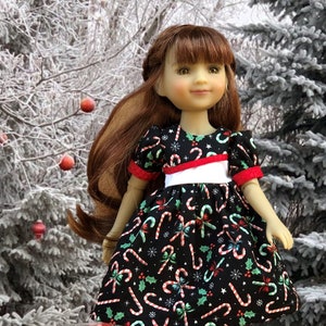 Puede incluir: Una muñeca con un vestido negro con un patrón de bastón de caramelo rojo y blanco. El vestido tiene una faja blanca y ribetes rojos. La muñeca tiene el pelo largo y castaño y lleva zapatos plateados.