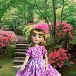 Op de afbeelding: Een pop die een roze en paarse bloemenjurk draagt met een witte kantversiering. De pop heeft blond haar en blauwe ogen. De pop staat voor een tuin met een stenen pad.