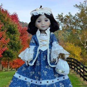 Puede incluir: Muñeca de porcelana con un vestido azul floral con ribete de encaje blanco y un gorro a juego. La muñeca tiene el pelo oscuro y sostiene un pequeño bolso blanco. El fondo presenta un colorido paisaje otoñal.