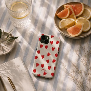 Op de afbeelding: Een wit smartphonehoesje met een herhalend patroon van kleine rode hartjes en het woord pommi in rode schrijfletters onderaan. Het hoesje heeft een uitsparing voor een dubbele camera aan de achterkant en een gladde, matte afwerking.