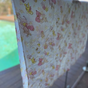 May include: A white baby blanket featuring a repeating pattern of pink and yellow cartoon animals, such as bunnies, bears, and ducks, and ABC blocks. The blanket is hanging on a clothesline outdoors.