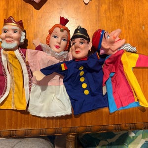May include: Four hand puppets featuring a king with a crown, a queen with a red crown, a soldier in a blue uniform with yellow buttons, and a jester in a red, yellow, and blue outfit. Each puppet has a moulded face and fabric clothing.
