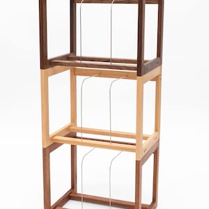 May include: A three-tiered wooden storage rack with a minimalist design. The rack is constructed from dark brown, light brown, and natural wood tones. It features three levels with metal rods for holding items. The rack stands on a white background.
