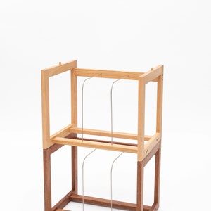 May include: A two-tiered wooden display stand with a light brown top section and a darker brown base. Thin, curved metal rods are suspended between the tiers. The stand is set against a white background.