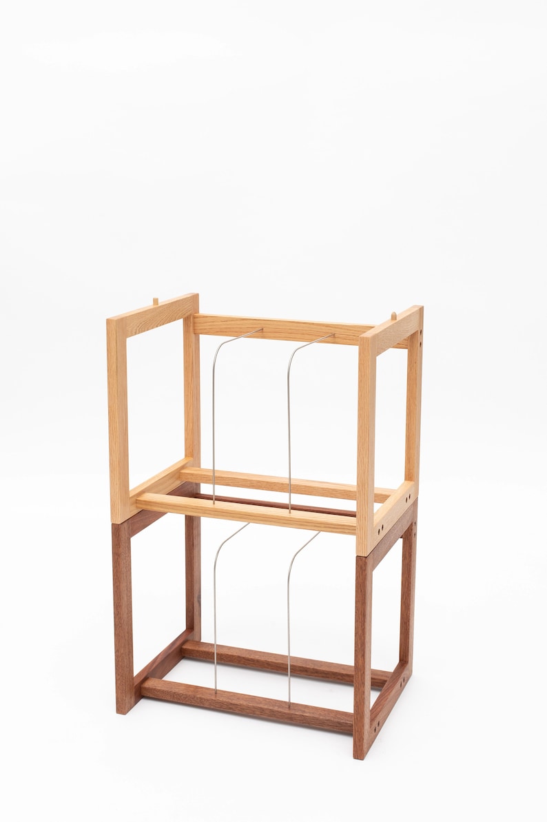 May include: A two-tiered wooden display stand with a light brown top section and a darker brown base. Thin, curved metal rods are suspended between the tiers. The stand is set against a white background.