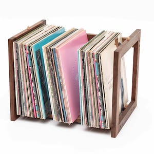 May include: A wooden record holder with a rectangular frame, holding a collection of vinyl records. The records have colorful album covers, including shades of blue and pink. The holder is made of dark wood.