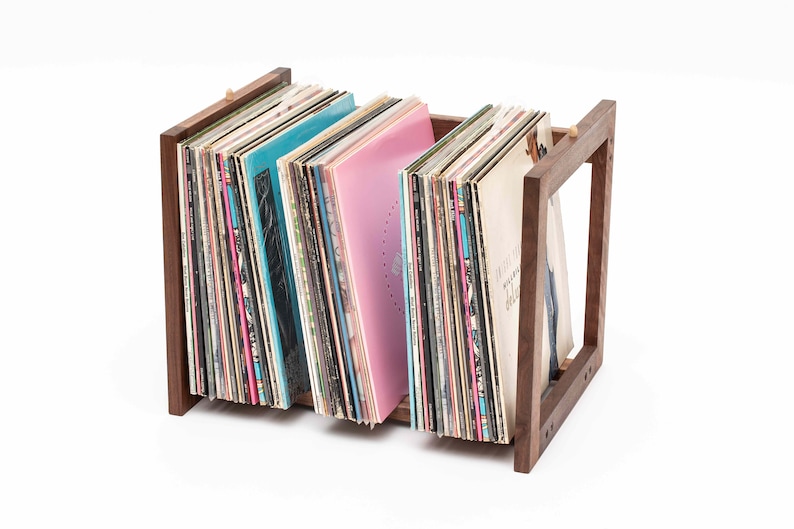 May include: A wooden record holder with a rectangular frame, holding a collection of vinyl records. The records have colorful album covers, including shades of blue and pink. The holder is made of dark wood.