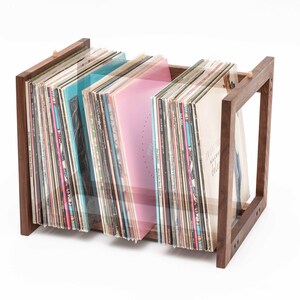 May include: A wooden record holder with a rectangular frame, holding a collection of vinyl records. The records have colorful album covers, including shades of blue and pink. The holder is made of dark wood.