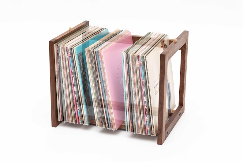 May include: A wooden record holder with a rectangular frame, holding a collection of vinyl records. The records have colorful album covers, including shades of blue and pink. The holder is made of dark wood.