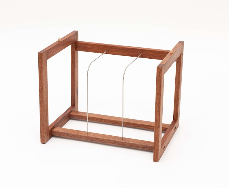 May include: A wooden bread slicer with a rectangular frame and three stainless steel wires. The wood is a warm brown color, and the wires are curved. The slicer is designed to cut bread evenly.