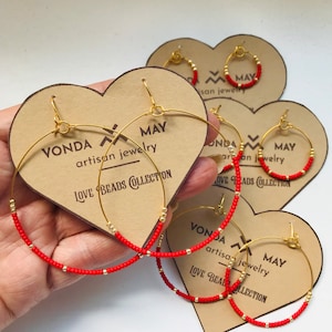 May include: Three pairs of gold hoop earrings with red beads. The earrings are displayed on heart-shaped cards with the text "VONDA MAY artisan jewelry LOVE BEADS COLLECTION".