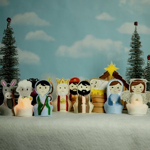 May include: A felt nativity set with finger puppets of Mary, Joseph, baby Jesus, an angel, a shepherd, a donkey, a sheep, and the three wise men. The puppets are arranged in front of a felt stable with a star on top. The set is on a white fluffy surface with two small silver Christmas trees in the background.