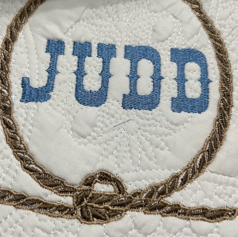 Może przedstawiać: Biały materiał z brązowym wzorem liny i wyhaftowanym imieniem "JUDD" w kolorze niebieskim.