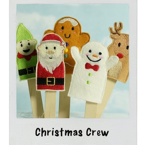 May include: Five felt finger puppets on wooden sticks. The puppets are a green elf, a red and white Santa Claus, a gingerbread man, a white snowman, and a brown reindeer. The puppets are arranged in a row and the text "Christmas Crew" is below them.