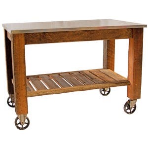 May include: A rustic kitchen island with a stainless steel top and a lower shelf made of wooden planks. The island is constructed from reclaimed wood and features four antique-style caster wheels for mobility. The overall color is a warm brown.