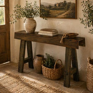 Puede incluir: Una consola de madera rústica con acabado oscuro, con un jarrón de cerámica envejecida con vegetación, libros y una cesta de mimbre. La mesa se apoya en patas anguladas, creando una estética de granja. Una pintura de paisaje enmarcada cuelga de la pared.
