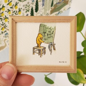 May include: A framed illustration of Winnie the Pooh standing on a stool and writing on a chalkboard. The chalkboard is green and has the numbers 1, 2, and 3 written on it. The illustration is signed "H. Shepard 36".