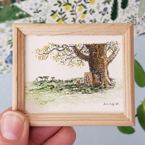 May include: A miniature watercolor painting in a light wood frame. The artwork depicts a large tree with a brown trunk and yellow leaves, with a small structure at its base. The scene is set on a green grassy hill.