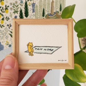 May include: A framed illustration of a yellow bear standing on a rug with the text "TAM HTAB" written on it.