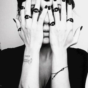 Matte black 3d printed eye ring displayed on a tattooed model's hands with long black nails covering her face