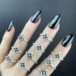 May include: A set of silver rings with black star-shaped accents. The rings are stacked on a hand with black nail polish.