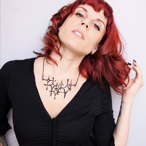 Matte black mid-century modern atomic sparkle necklace displayed on a tattooed model with red hair wearing a low cut black dress. Witchy 3d printed jewelry by Salem, MA artist Hypnovamp.