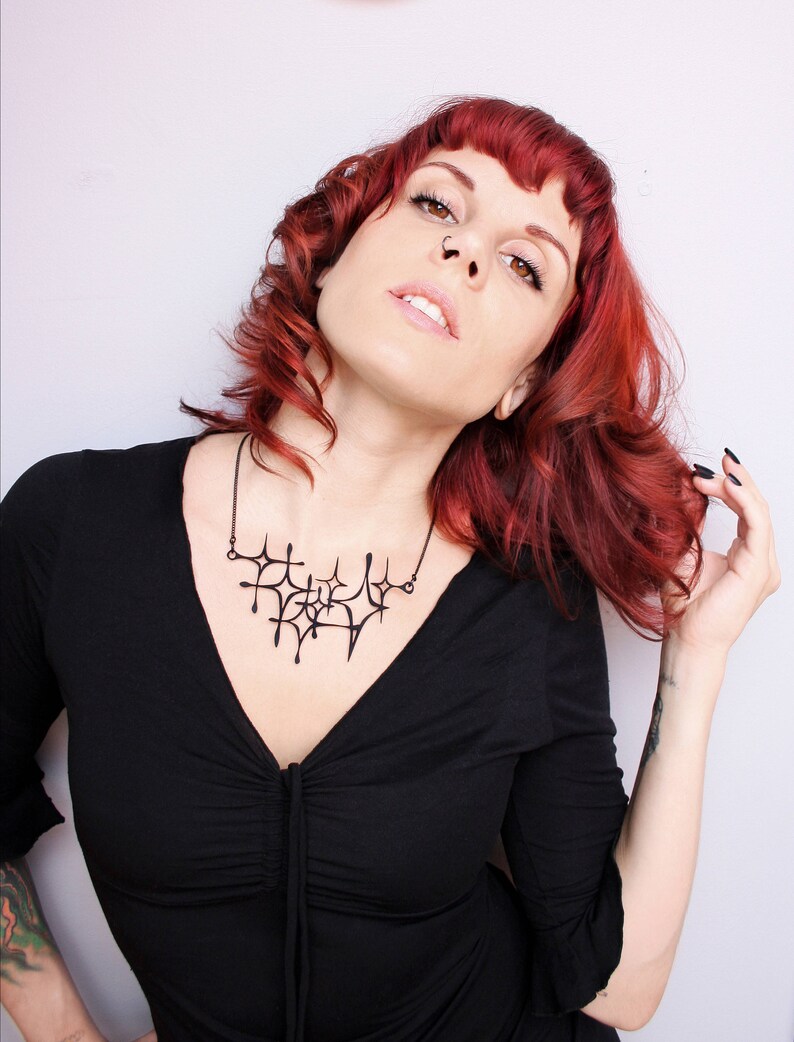 Matte black mid-century modern atomic sparkle necklace displayed on a tattooed model with red hair wearing a low cut black dress. Witchy 3d printed jewelry by Salem, MA artist Hypnovamp.